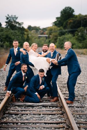 Wedding party creates fun group pose on railroad tracks wearing navy suits.