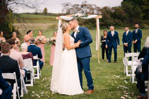 Outdoor wedding ceremony takes place under wooden arch with guests seated on white chairs in grassy field.