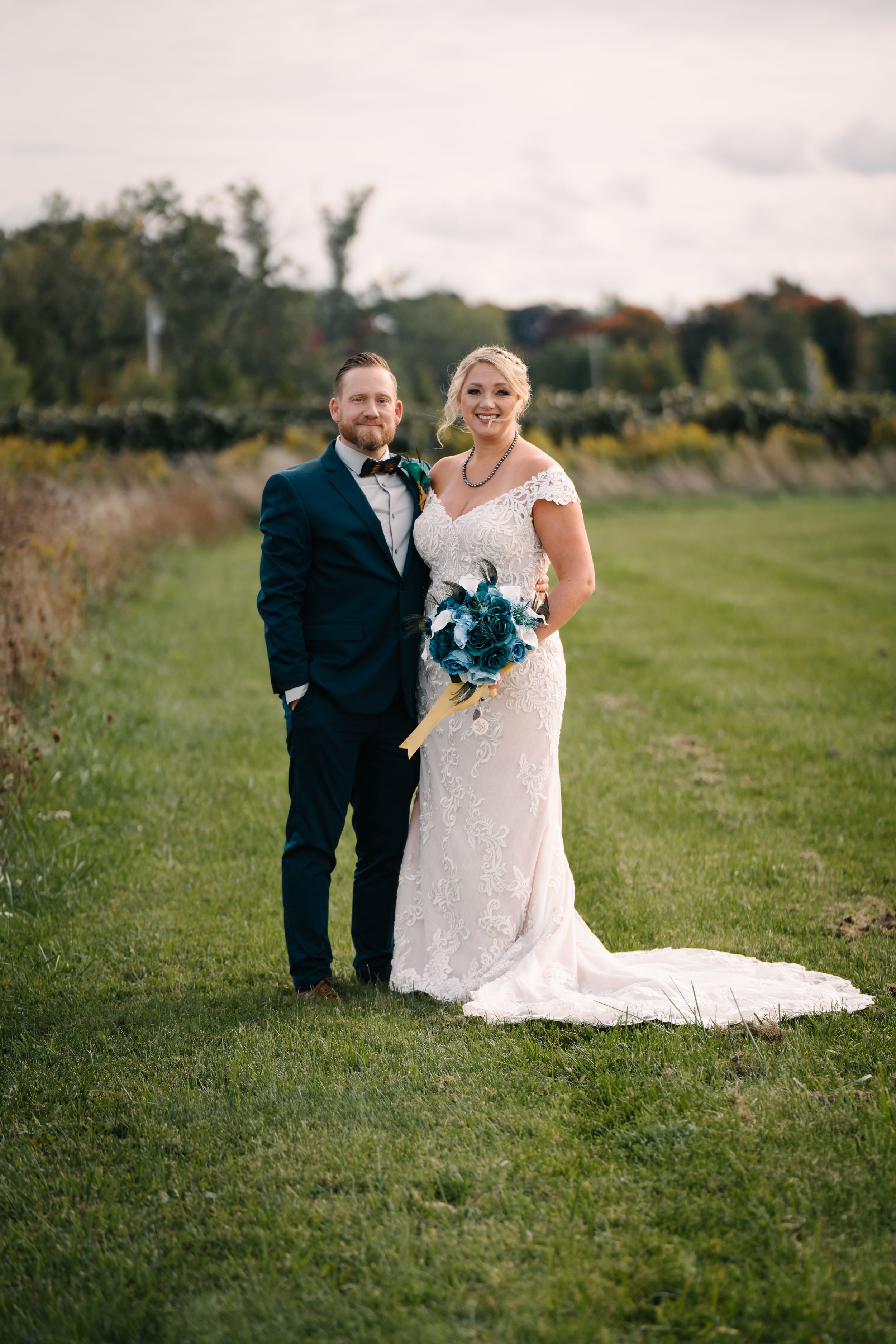 Newlywed couple posing in countryside setting with bride in lace gown holding blue bouquet.