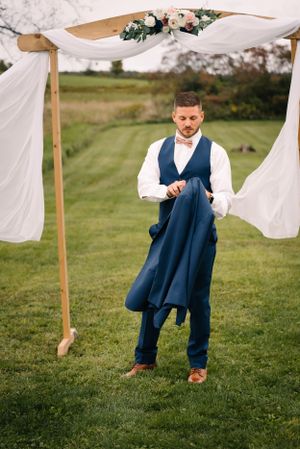 Groomsman putting on a navy blue suit jacket under a floral wedding arch with white draping.