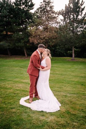 A couple shares a romantic moment on a lawn with pine trees in the background, wearing rust-colored suit and white gown.