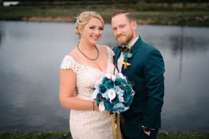 Wedding couple stands near a lake with teal floral bouquet and navy suit on an overcast day.