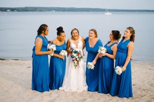 Bride and bridesmaids in blue dresses share candid moment on beach.