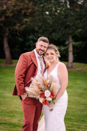 Wedding couple poses outdoors in burgundy suit and white dress with fall bouquet.