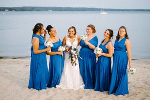 Bridal party in vibrant blue dresses laughs together on beachfront.