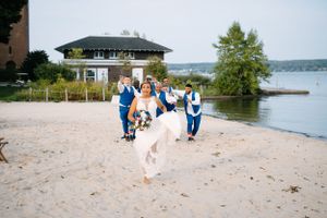 Wedding party runs joyfully on a sandy beach with a gazebo and lake in the background.