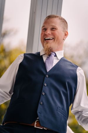 A joyful groom in a navy vest and purple tie laughs during an outdoor wedding portrait.