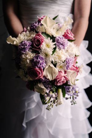 An elegant bridal bouquet features pink roses, white calla lilies, and purple flowers against a white dress.