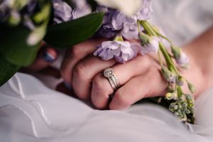 Close-up detail of wedding rings and delicate purple flowers against white fabric.