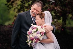An emotional wedding couple embraces while holding a pink and purple bouquet.
