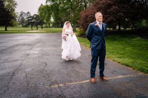 A bride and groom stand separately during their first look on a wet driveway.
