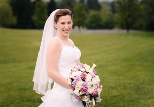 A bride smiles radiantly while holding her pink and purple bouquet outdoors.