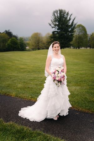 A bride poses on a paved path holding a pink and purple bouquet against a green landscape.