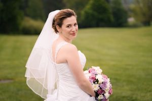A bride in a layered white gown poses with her bouquet against a green landscape.