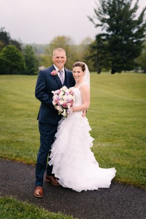 Wedding couple poses outdoors on path with pink bouquet against green lawn and evergreen trees.