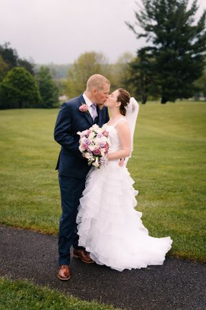 Wedding couple shares intimate moment on pathway with pink floral bouquet and scenic backdrop.