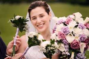 Bride smiles while holding pink and white floral bouquet outdoors.