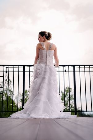 Back view of bride in layered wedding gown standing against iron railing with scenic view.