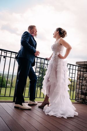 A couple in wedding attire share a playful moment on a scenic deck overlooking a green landscape.