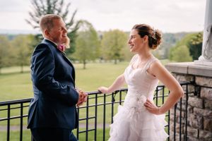 Wedding couple laughs together on a stone-railed balcony with rolling hills in the background.