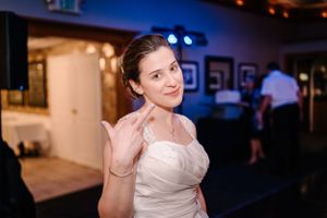 A bride in white gestures playfully at an indoor reception venue with blue lighting.