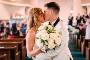 Romantic kiss between newlyweds holding a white rose bouquet in a church setting.