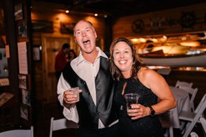 Wedding guests celebrate with wine glasses at a rustic indoor reception venue with warm lighting.