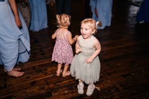 Two small children in dresses dance together on a wooden floor at a wedding reception.