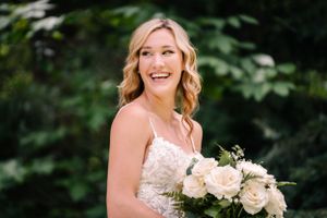 The bride smiles while holding a lush bouquet of cream roses against a green forest backdrop.