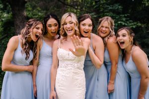 Bridesmaids in light blue dresses surround bride in white, all showing excited expressions.