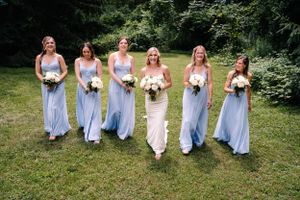 Bridal party poses in matching light blue dresses with white bouquets in outdoor garden setting.