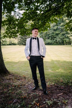 A person in formal attire poses outdoors under a tree wearing a white jacket and black bow tie for a special occasion.