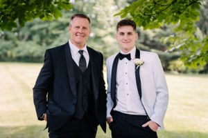 Two people in tuxedos and boutonnieres pose together under green trees for a formal outdoor portrait.