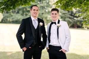 Two formally dressed people smile while posing together outdoors in black and white suits with boutonnieres.