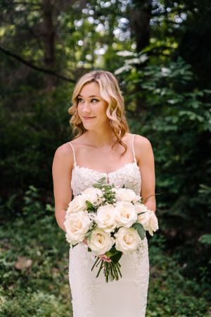 A bride holds a fresh bouquet of cream roses against a wooded backdrop.