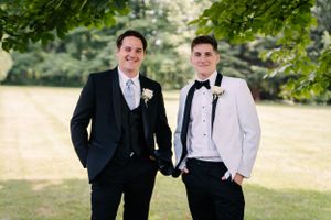 Two people in contrasting formal suits stand together for a portrait under green tree branches.