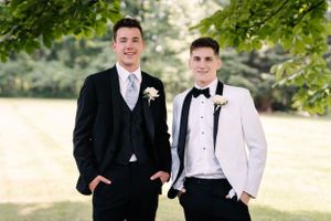 Two people smile while posing in formal attire with boutonnieres under tree branches outdoors.