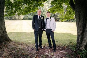 Two people in formal attire stand between trees for an outdoor black and white portrait.
