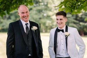 Two people in formal suits and boutonnieres share a candid moment during an outdoor photo session.