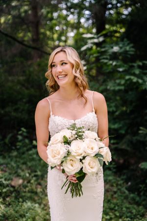 A bride in a delicate lace dress holds a classic bouquet of cream roses in a forest setting.
