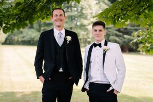 Two people in contrasting formal wear pose casually under trees for an outdoor portrait.