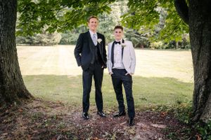 Two people pose in formal suits for a black and white outdoor portrait between large trees.