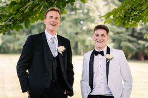 Two groomsmen pose together outdoors, one in black suit and one in white tuxedo jacket.