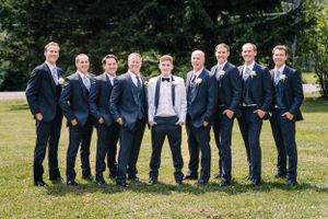 Wedding party stands in a row outdoors wearing navy suits with one member in white standing in the center.