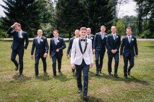 Fun wedding party photo with groomsmen jumping in navy suits behind central figure in white tuxedo.