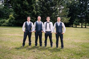 Four members of wedding party in vests stand on grass with evergreen trees in background.