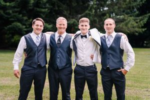 Four groomsmen pose casually in matching vests and dress shirts against evergreen backdrop.