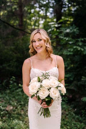 A bride in white lace holds a classic bouquet of cream roses against forest greenery.