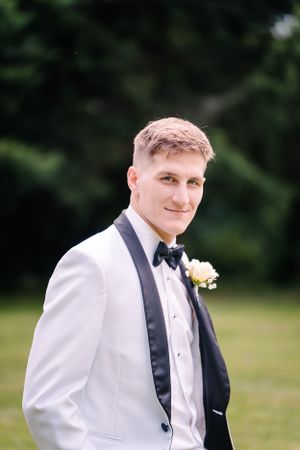 Formal portrait of groom in white tuxedo jacket with black bow tie against natural background.