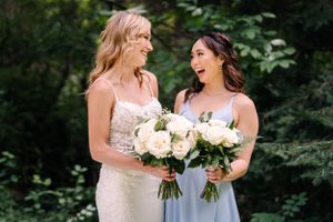Two wedding attendants share a joyful moment while holding matching cream rose bouquets.